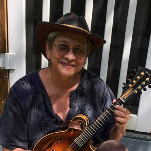 Debra Amesqua wearing a black cowboy hat and a toothy smile holds a small guitar as though she's about to start playing.
