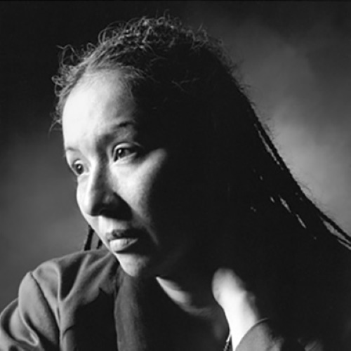 In this atmospheric black and white photo, Ingrid Washinawatok looks down and off to the left away from the camera, her hair in main thin braids draped over her shoulders, and hand placed on her neck.