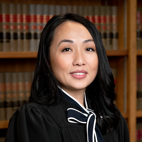 In this formal portrait of Kristy Yang, she is seated in front of a bookshevles of law books. She smiles politely at the camera, her black hair draped across her shoulders. She wears a black shirt with a white edged black bow tied at the collar and her judge robes.