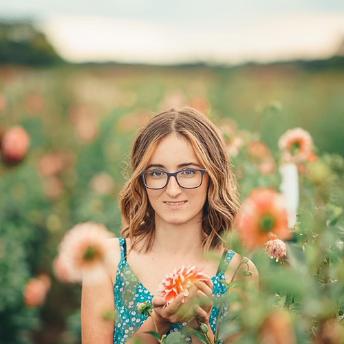 Madelyn, a short brown haired young woman wearing glasses, stands in a field of flowers wearing a teal floral tank dress. She holds one bloom and smiles slightly at the camera.