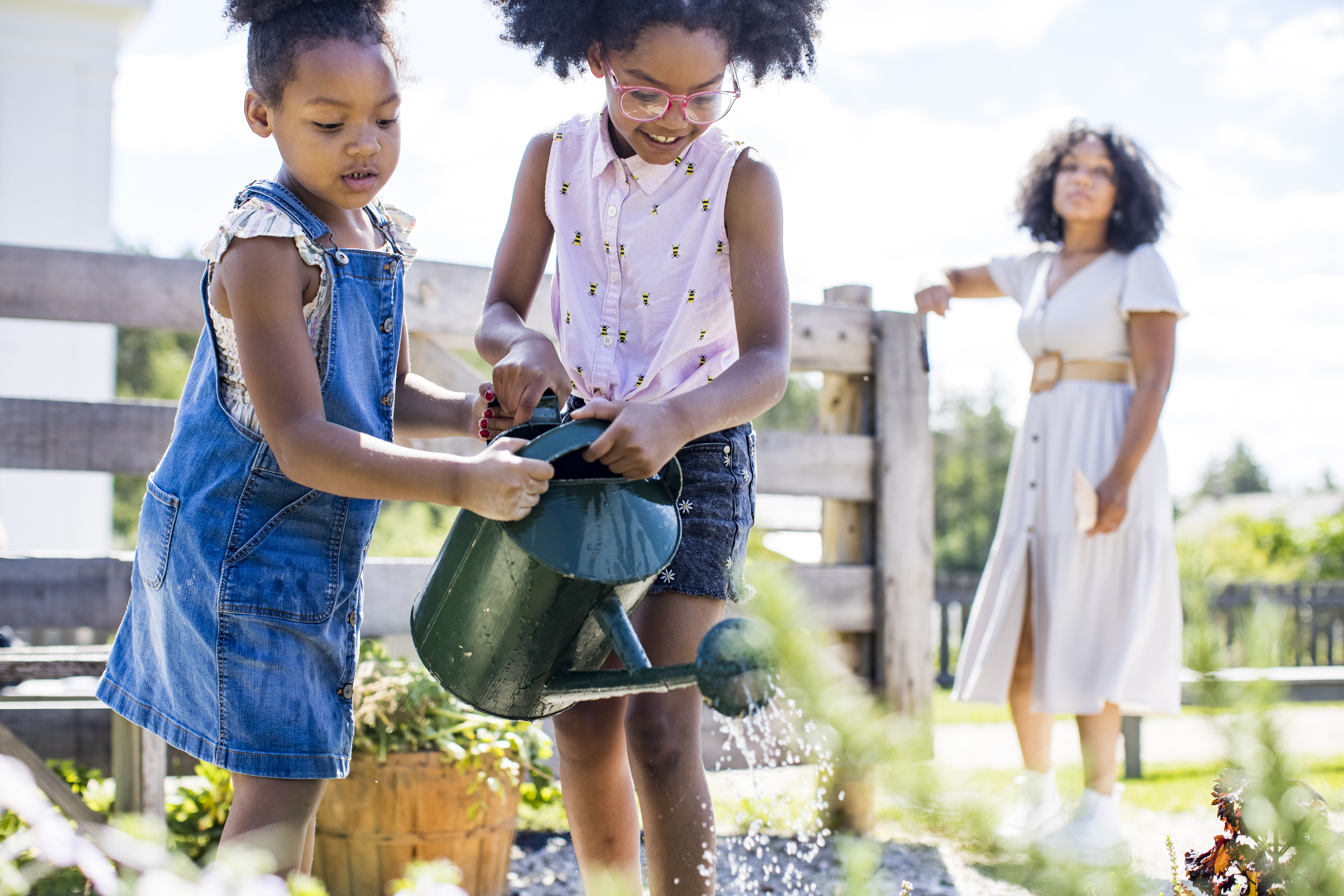 Two young black girls water flowers while their mother looks on to help them.