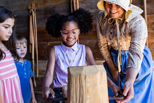 A young black girl with cute hair poms smiles excitedly as she's taught how to work with wood by a white woman in period clothing, as some children and a man look on with curiosity.