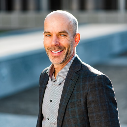 A man with a shaved head wearing a plaid shirt and blazer smiles at the camera