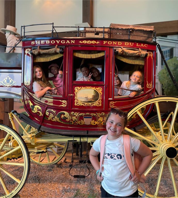 A group of children inside of a red and yellow carriage with a young girl standing outside and posing for a picture at Wade House 
