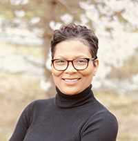 A color photo of a smiling black woman with curly hair. She is wearing glasses.
