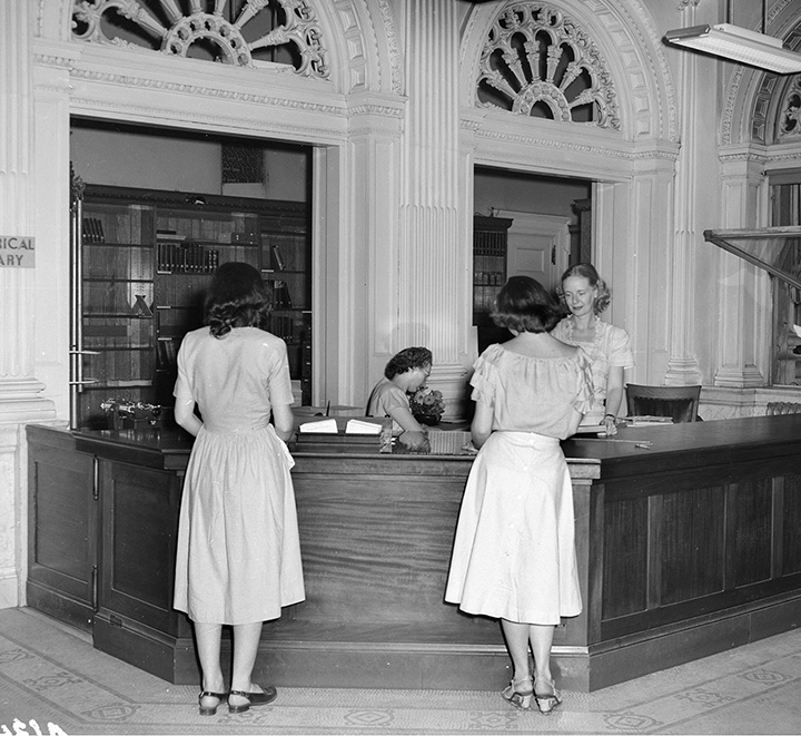 Black and white image of two women at the Wisconsin Historical Society's 2nd floor library circulation desk while one staff member answers a phone and another assists them.