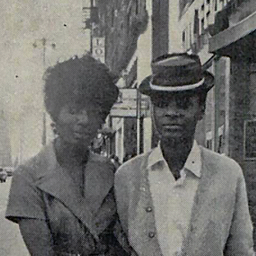 Manonia Evans (left) and Donna Burkett, 1971. Manonia Evans wears a wide labeled dress with short almost capped sleeves, her hair is curly and short, a small smile plays across her lips. Donna Burkett wearing a pork pie hat with a light ribbon, set at a jaunty angle and casual light suit. They stand close together with Evans' arm wrapped around one of Burkett's.