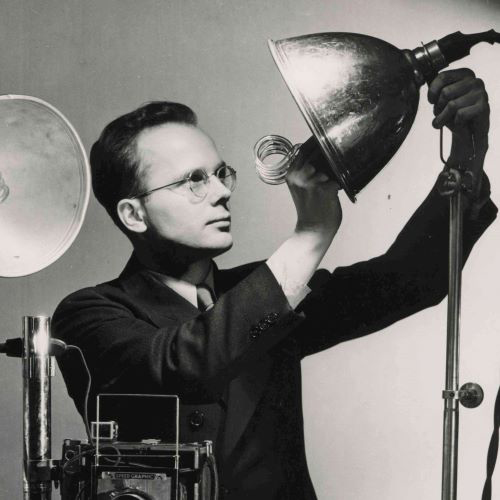 Edwin Farber examines a large light fixture looking focused and wearing a smart suit and glasses.