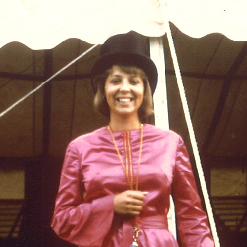 Elizabeth Fentress wears a bright magenta floor length satin gown. She smiles proudly in her top hat directly facing the camera in front of the big top.