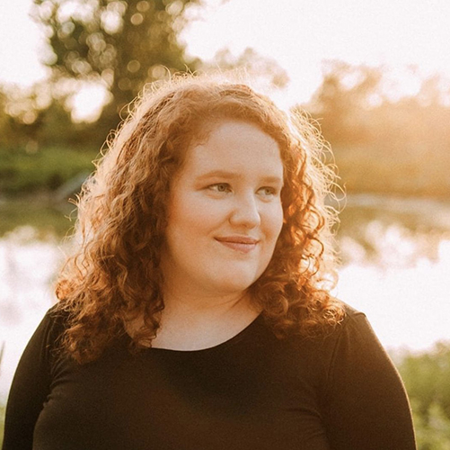 Kaia, a curly haired young woman, stands near a pond looking off to the side with the glow of the sun haloing her hair and shoulders. She has a slight smile and a black scoop neck shirt on.