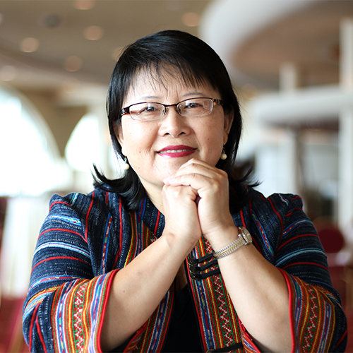 A formal headshot of Mai zong Vue, smiling at the camera, wearing glasses and a multi-color woven quarter length sleeve jacket. Her hands are clasped in front of her and she rests her cheek on her fingers. Her dark hair is partially pulled back and her bangs, dust her glasses.