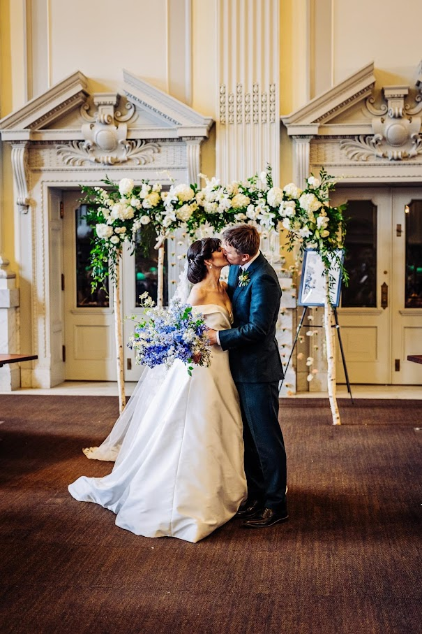 A couple are photographed under their wedding arch in the library kissing at the end of their ceremony