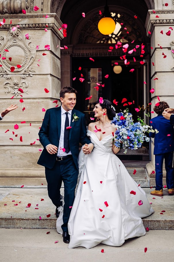 A smiling hetero couple, walks out onto the terrace with flower petals floating around them in pinks and reds. The bride wears and off shoulder low neckline princess cut gown, the groom a navy suit. Both are smiling excitedly as they hold hands.