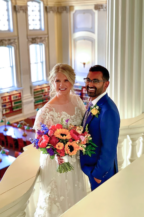 A blonde bride and a dark haired groom, smile delightedly on the balcony overlooking the library. They are holding a large colorful sunflower boquet between them.