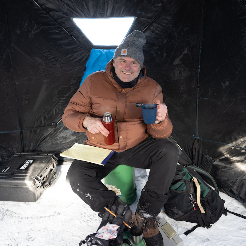 Jim smiles excitedly from inside a tent, bundled for winter and drinking his coffee.