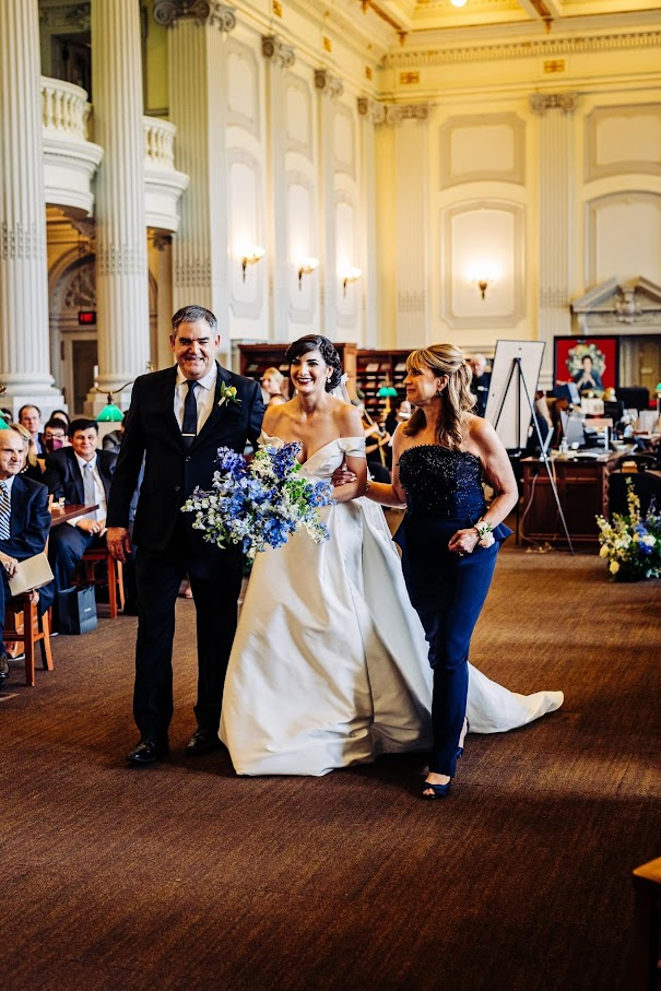 A bride walks excitedly downt the isle, flanked by her parents headed towards the alter.