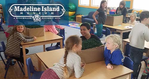 Madeline Island Museum. Kids work on multiple grouped desks in a classroom, some smile at the camera.