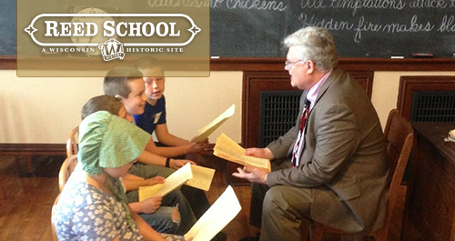 Reed School. A group of kids engage animatedly with an older man instructing them from a one room school house desk.