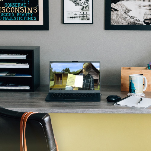 A desk with a laptop showing the Wiscosin History Website, surrounded by a notebook, mug, file shelf, and some Wisconsin themed art.