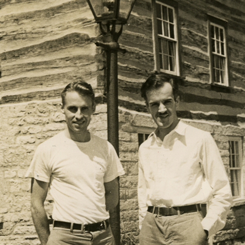 Bob Neal and Edgar Hellum stand in fron of the Pendarvis historic building by an iconic street lamp. They stand close but not touching, both in somewhat casual shirts and pants, slightly smiling on this sunny day in this sepia photograph.