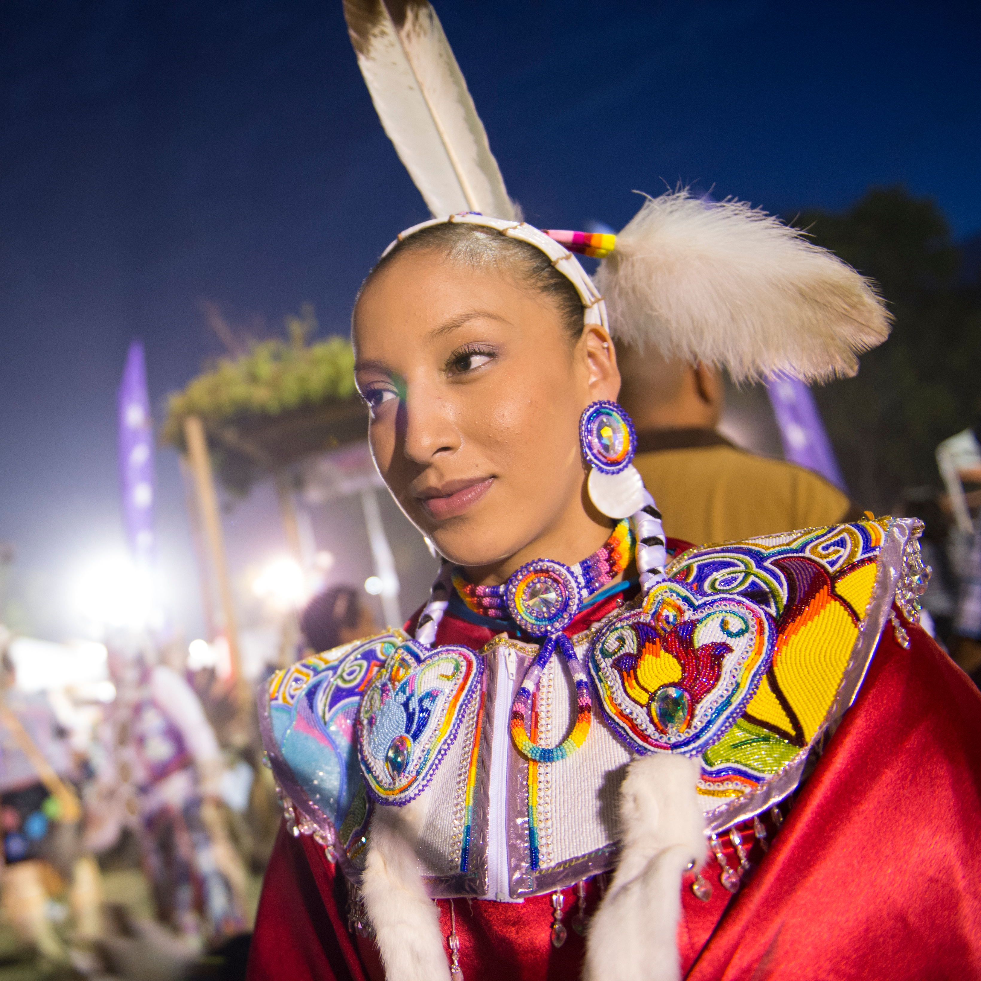 A woman dressed in red and rainbow regalia with many beaded floral pieces and beaded collar, stands proud in the foreground of a croud blurry behind her. She has a long beaded headband with feathers on her head, her hair braided tightly with two wrapped braids down her front. 