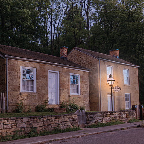 Pendarvis house at dusk,  orange colored brick and white trim windows and doors. A stone wall lines the house next to the road. The lamp is lit. 