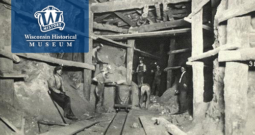 Wisconsin Historical Museum. Workers stand and sit around in a mine under ground in this black and white image.
