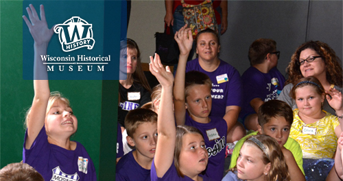 Wisconsin-Historical Museum. A large group of students and chaperones wearing matching shirts animatedly engage with a speaker off camera. Several girls raise their hands high!